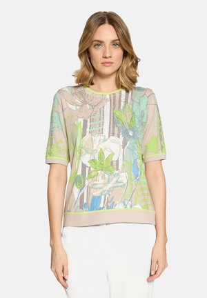 Femme aux cheveux blonds mi-longs portant un haut beige à manches courtes avec des motifs floraux et rayés verts, bleus et blancs, associé à un pantalon blanc.