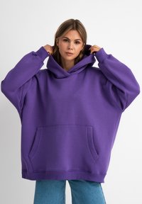 Felpa viola oversize con tasca frontale, polsini a costine e un grande cappuccio, realizzata in morbido materiale, abbinata a jeans azzurri chiari.