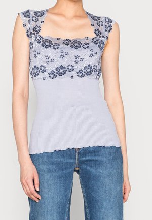 Kobieta w lawendowym bezrękawnym prążkowanym topie z kwiatową koronkową dekoracją i niebieskich dżinsach, stojąca na jednolitym tle.