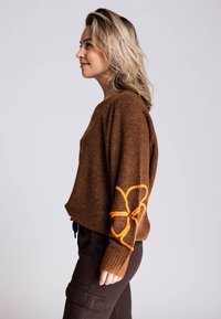 Brauner Strickpullover mit gerippten Bündchen und einem orange bestickten Blumendetail am Ärmel. Wird mit schwarzen Cargohosen getragen.