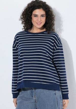 Vrouw met krullend haar, gekleed in een marineblauw-wit gestreept shirt met lange mouwen en lichtblauwe jeans, glimlacht zachtjes terwijl ze voor een witte muur staat.