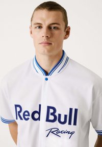 Witte trui met blauwe accenten en een knoopsluiting. Voorzien van het "Red Bull Racing" logo in marineblauw op de voorkant, met een gestreepte ribbelkraag.