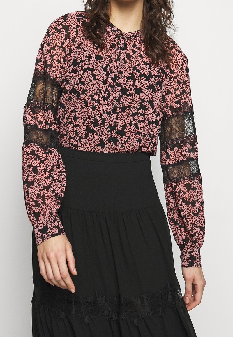 Blusa negra con patrón floral rosa, mangas de encaje traslúcidas, cuello redondo y detalles de botones. Presenta un diseño fruncido en los hombros.