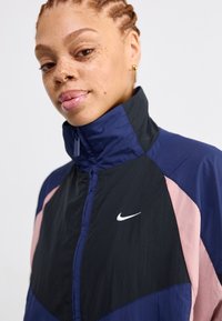 Giacca Nike con colletto alto, design a blocchi di colore in nero, blu navy e rosa, con un logo bianco sul petto. Tessuto strutturato.