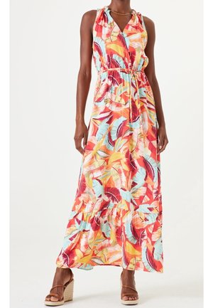 Femme portant une robe maxi sans manches à imprimé tropical coloré de feuilles, taille cintrée, et des sandales compensées marron sur fond blanc.