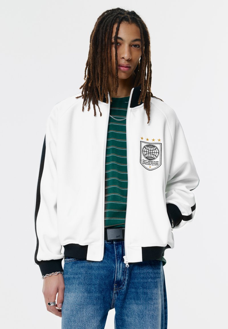 Ung person med lange dreadlocks iført en hvid zip-up jakke med sorte striber og et globus emblem, grønstribet skjorte og blå jeans.