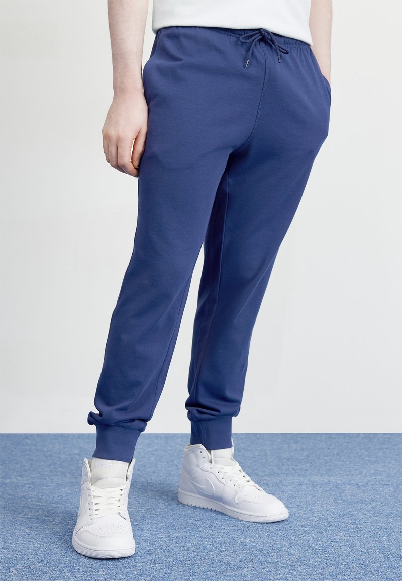 Marineblauwe, zachte katoenen joggingbroek met elastische tailleband en boorden. Voorzien van zijzakken en een trekkoordsluiting. Draagt wit sneakers.