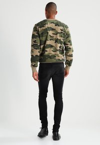 Felpa camouflage in verde, beige e nero, con polsini e orlo a coste; abbinata a jeans neri slim e scarpe nere con lacci.