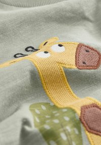 Light green fabric features an embroidered yellow giraffe. Giraffe has black accents and a brown spotted patch, against a green leaf design.