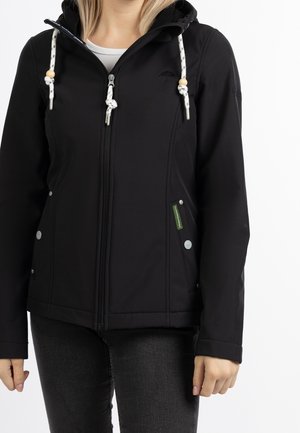 Femme portant une veste à capuche noire avec des cordons blancs et des perles en bois, fermée par une fermeture éclair, associée à un jean gris foncé sur un fond blanc.