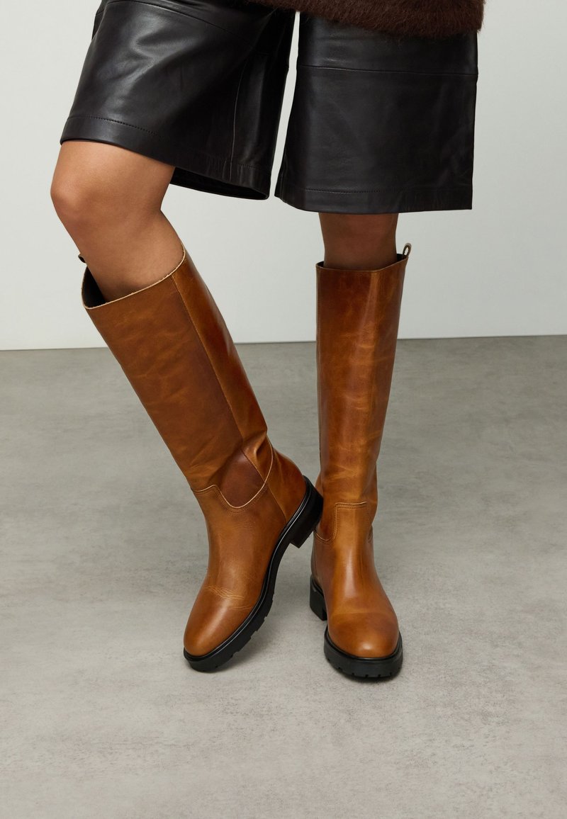 Bruine leren kniehoge laarzen met een gladde afwerking, een afgeronde neus en een zwarte rubberen zool. Voorzien van een uitsnijding aan de zijkant voor extra design.