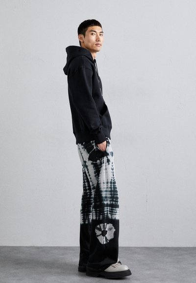 Schwarzer Hoodie mit einer Fronttasche, kombiniert mit schwarz-weißen Batik-Hosen mit floralem Muster. Das Model steht vor einem neutralen Hintergrund.