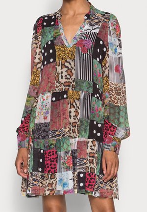 Pitkähihainen tilkkuhaalari, jossa on sekoitettu eläinkuoseja, pilkkuja, raitoja ja kukkakuvioita sekä V-pääntie, päällä seisovalla henkilöllä.