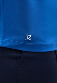 Γαλάζιο τελείωμα πουκάμισου με μικρό λευκό σχέδιο σε σχήμα καρδιάς πάνω από τη μέση σκούρου παντελονιού.