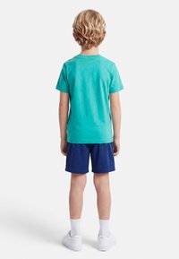 Ξανθό παιδί που στέκεται με την πλάτη, φοράει τιρκουάζ μπλουζάκι, σκούρο μπλε σορτς, λευκές κάλτσες και λευκά αθλητικά Puma σε μονόχρωμο φόντο.