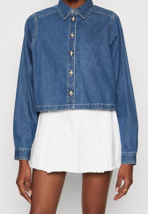 Woman wearing a cropped blue denim button-up shirt with long sleeves over a white pleated mini skirt, standing against a plain background.