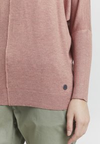 Maglione rosa chiaro realizzato in tessuto morbido, caratterizzato da un orlo a coste, una vestibilità leggermente oversize e un'accenno di logo nero rotondo di lato.
