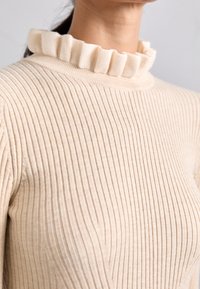 Maglione beige a coste con un collo alto e arricciato. La texture è morbida e il tessuto sembra elastico, evidenziando una silhouette aderente.
