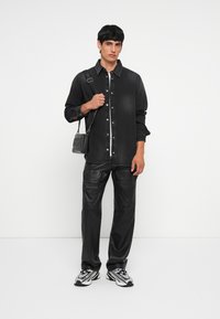 Chemise en denim noir à boutons avec des rayures verticales. Pantalons en cuir noir et baskets noires avec des accents en argent. Portant un sac bandoulière noir.