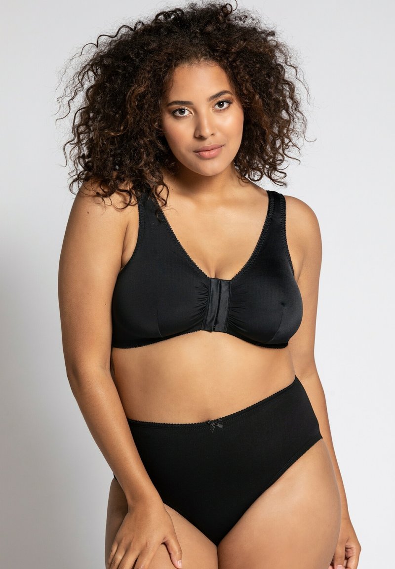 Schwarzer kabelloser BH mit Frontverschluss, aus weichem Stoff, breiten Trägern und dezenten Nähten. Hochgeschnittene schwarze Panty mit Spitzenakzenten.