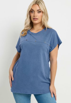 Blondynka w luźnym niebieskim topie z krótkim rękawem i guzikami na piersi oraz w jasnoniebieskich dżinsach, stojąca na białym tle.