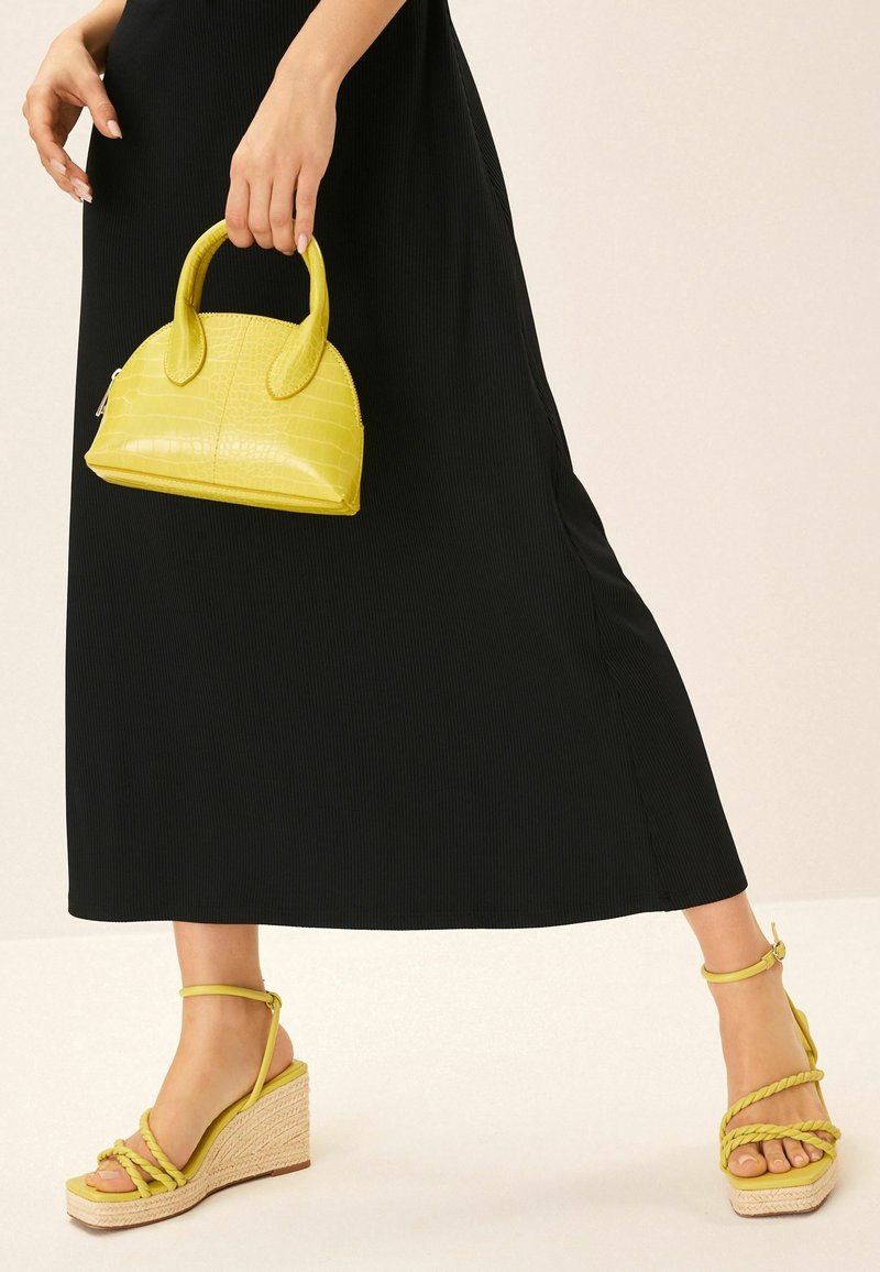 Femme en robe noire tenant un petit sac à main jaune, portant des sandales compensées jaunes avec des brides à la cheville et des détails tressés.