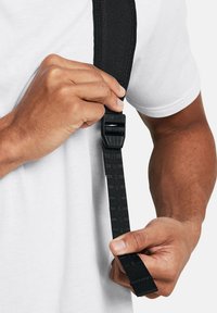 Sangle noire réglable en nylon texturé, avec une boucle en plastique. Une main agrippe la sangle, avec un t-shirt blanc uni visible en arrière-plan.