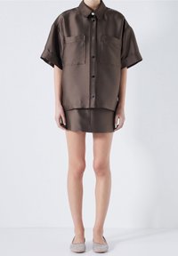 Brązowa satynowa koszula z krótkim rękawem z dwiema przednimi kieszeniami, zapięcie na guziki oraz dopasowana mini spódniczka. Miękka tekstura, luźny krój i minimalne zdobienia.