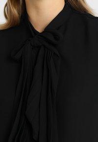 Svart blus med design för knytkrage, har draperat tyg och mjuk textur, utan synliga mönster eller detaljer i metall.