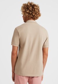 Beige polo skjorte med korte ærmer, der har en blød tekstur og en krave. Bagfra ses rene linjer og minimal detaljer.