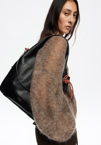 Czarny skórzany plecak ze srebrnymi elementami, z teksturowanym wykończeniem i czerwonymi ozdobnymi akcentami. Częściowo widoczny miękki brązowy sweter.