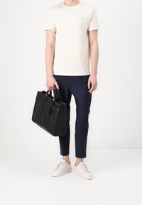 Czarna torba typu tote ze skórzanym materiałem, dwoma uchwytami, prostokątnym kształtem i gładką fakturą. Modelka nosi kremowy t-shirt, granatowe spodnie i białe trampki.