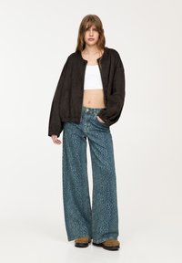 Svart bomberjacka med dragkedja framtill, kombinerad med vida blå jeans med ett subtilt leopardmönster. Modellen bär bruna träskor.