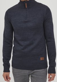 Pull en maille noir gris à demi-zip avec col, poignets et ourlet côtelés, porté avec un jean gris clair, montrant le torse avant et les bras.