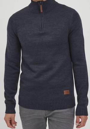 Suéter de punto gris oscuro con media cremallera y cuello, puños y dobladillo acanalados, combinado con jeans gris claro, mostrando la parte frontal del torso y los brazos.