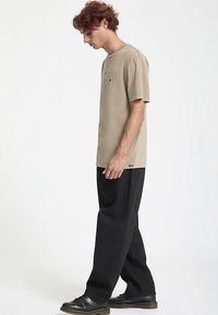 Tan short-sleeve t-shirt with a round neckline and small logo embroidery. Paired with loose-fitting black pants and black footwear.