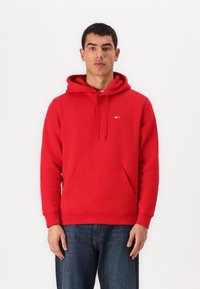 Felpa rossa in cotone con tasca a canguro, cappuccio regolabile con cordino e un piccolo logo sul petto. Polsini e orlo a coste.