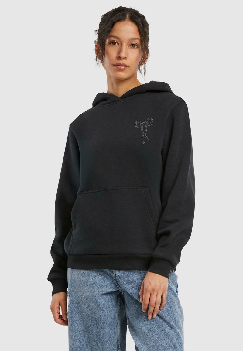 Svart hettegenser med lommer foran og en subtil sløyfegrafikk på venstre bryst. Brukt med lyseblå baggy jeans. Myk stofftekstur.