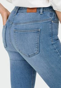 Zilās denima bikses ar vidējiem augstumiem, aizmugurējām kabatām ar nostiprinātu šuvēm un ādas logotipa uzšuvumu jostasvietā.