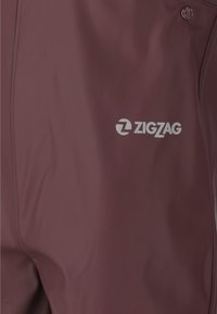 Pantalones impermeables color burdeos hechos de material sintético suave, con un logo "ZIGZAG" impreso en plata reflectante en el muslo.