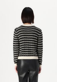 Osoba z krótkimi ciemnymi włosami, ubrana w czarno-białą pasiastą swetrową bluzę i czarne skórzane spodnie, stojąca plecami na białym tle.