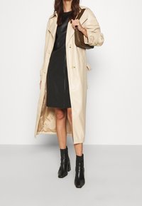 Beige Trenchcoat mit einer glatten, glänzenden Oberfläche, doppelreihigem Design und goldenen Knöpfen, kombiniert mit einem schwarzen, figurbetonten Kleid und schwarzen Stiefeletten.