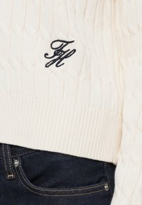 Ivory sweter w splot kablowy z żebrowanym dołem, z czarnym haftowanym logo. Połączony z ciemnoniebieskimi dżinsami, prezentującymi detal z metalowym guzikiem.