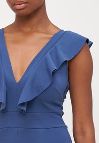 Μπλε φόρεμα με V-λαιμόκοψη, που διαθέτει φουσκωτές λεπτομέρειες στους ώμους και εφαρμοστό μπούστο. Απαλή υφή, μινιμαλιστικός σχεδιασμός.