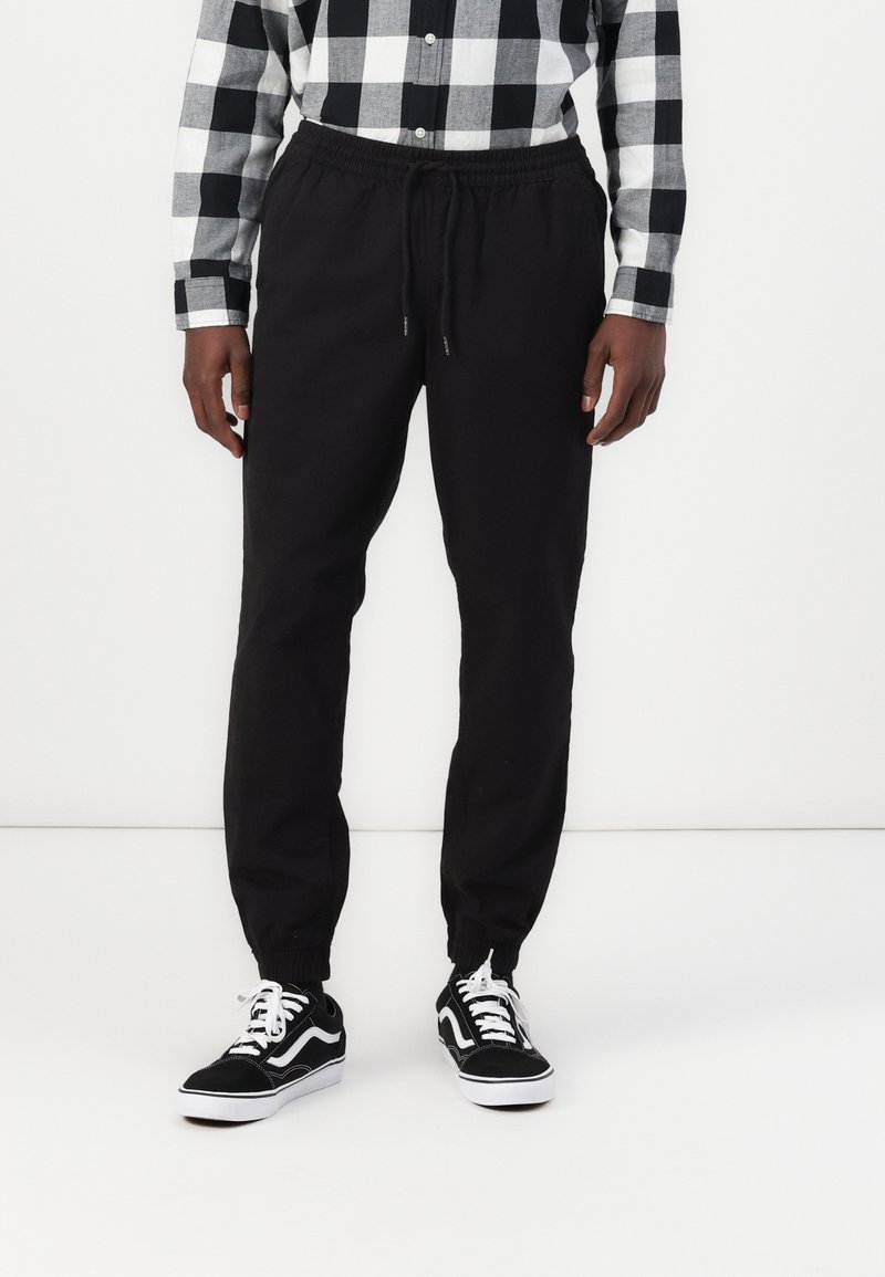 Zwarte sweatpants met elastische boorden en een tailleband met trekkoord, gecombineerd met zwart-witte sneakers. Model draagt een geruit shirt.