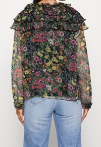 Blouse fleurie en tissu noir transparent avec des motifs floraux multicolores, un col à volants et des boutons sur les manches.