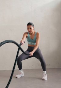 Fitness trening s ženom koja drži crni battle rope, obučena u svijetloplavu majicu bez rukava i crne tajice, u minimalističkom unutarnjem prostoru.