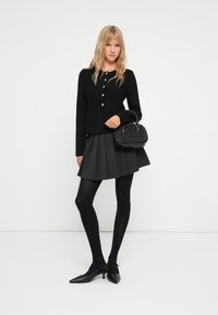 Cardigan noir avec boutons en argent, jupe plissée grise, collants noirs et escarpins noirs pointus. Petit sac à main texturé noir.