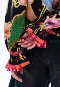 Ręce złożone za plecami, w kolorowej bluzce w kwiatowy wzór z falbaniastymi różowymi mankietami, zestawione z czarnymi dżinsami.