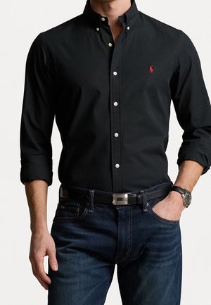 Man draagt een zwart overhemd met knopen en een rood logo, opgerolde mouwen, blauwe spijkerbroek, zwarte riem en polshorloge, hand in de zak.